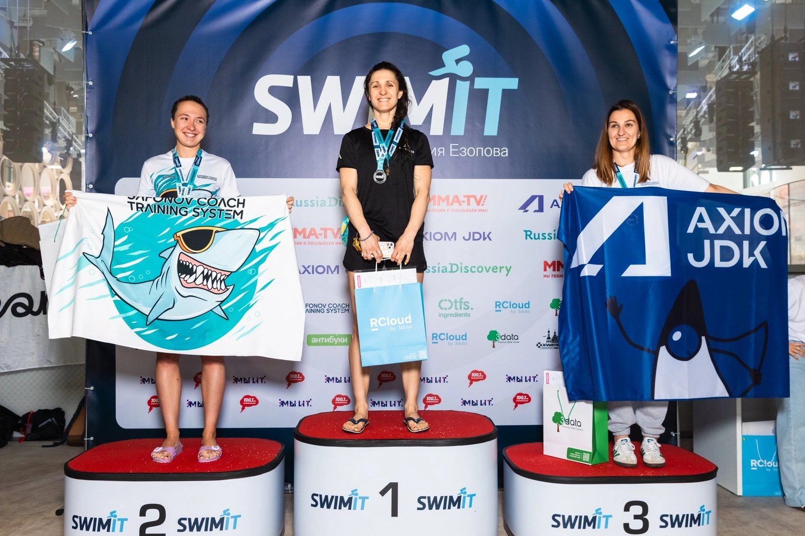 Три женщины стоят на призовом подиуме с медалями и рекламными флагами после соревнований по плаванию