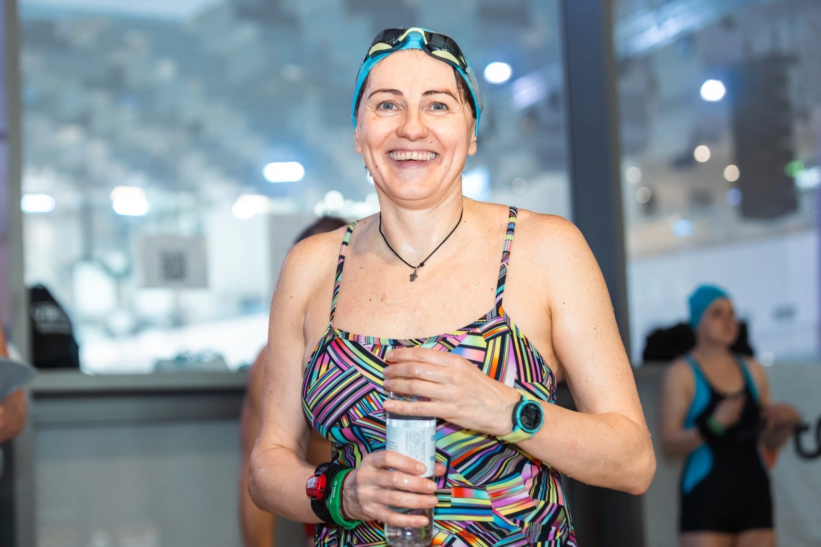 Улыбающаяся женщина в купальнике и плавательной шапочке держит бутылку воды у бассейна