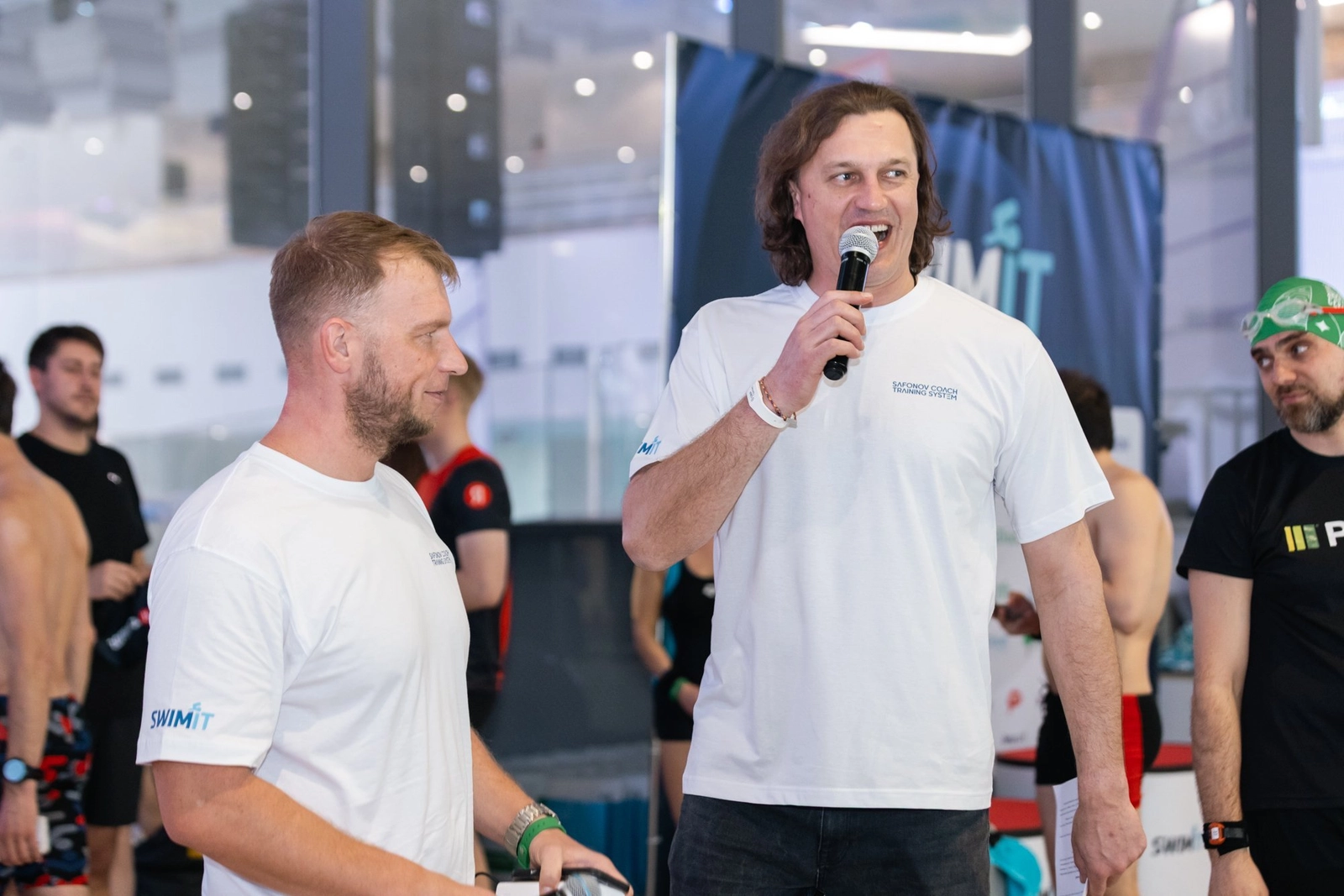 Виктор Набутов и Сергей Сафонов стоят у бассейна; Виктор говорит в микрофон, Сергей слушает