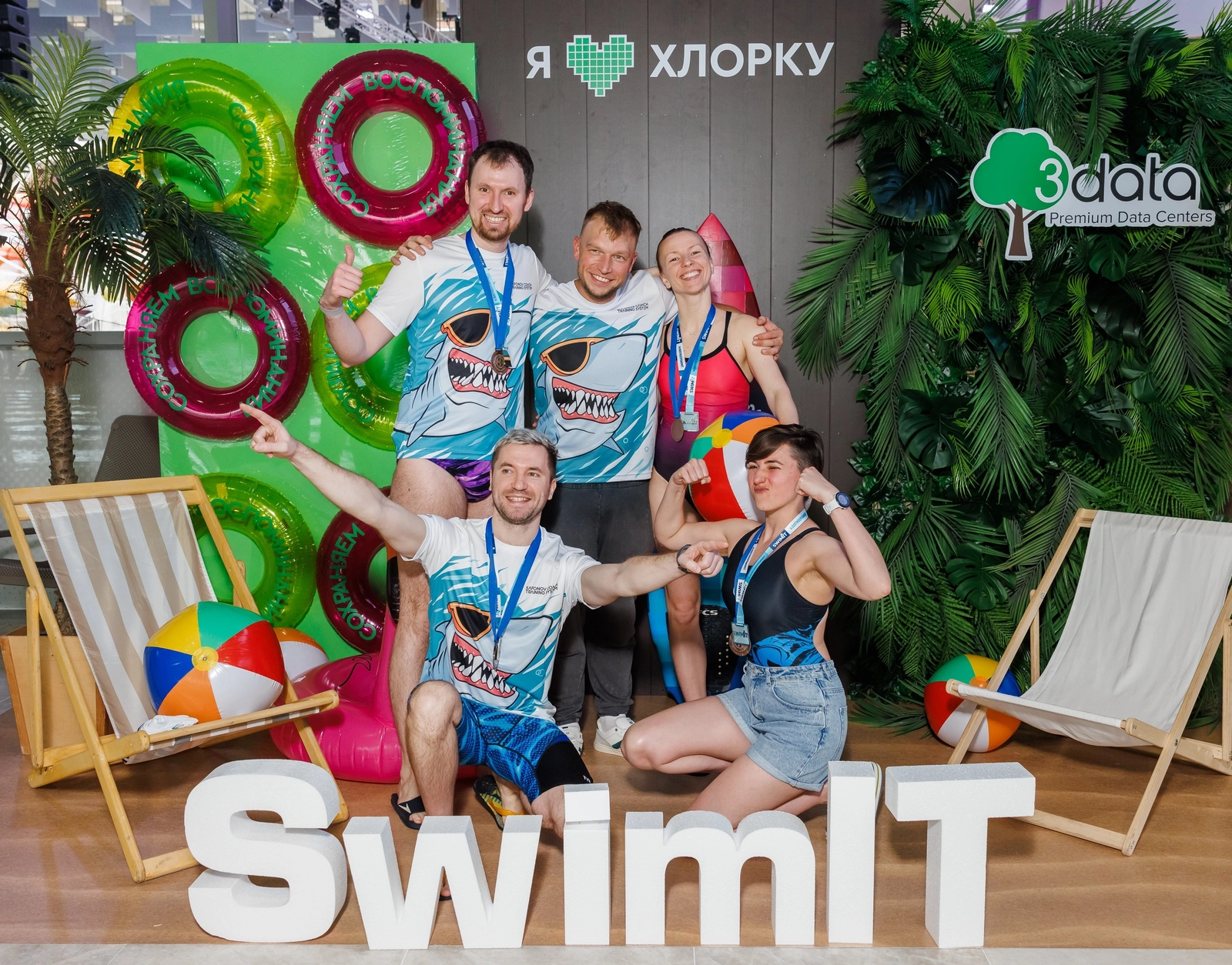 Группа из пяти человек позирует в фотозоне на тему плавания с надувными кругами, шезлонгами и надписью "SwimIT"